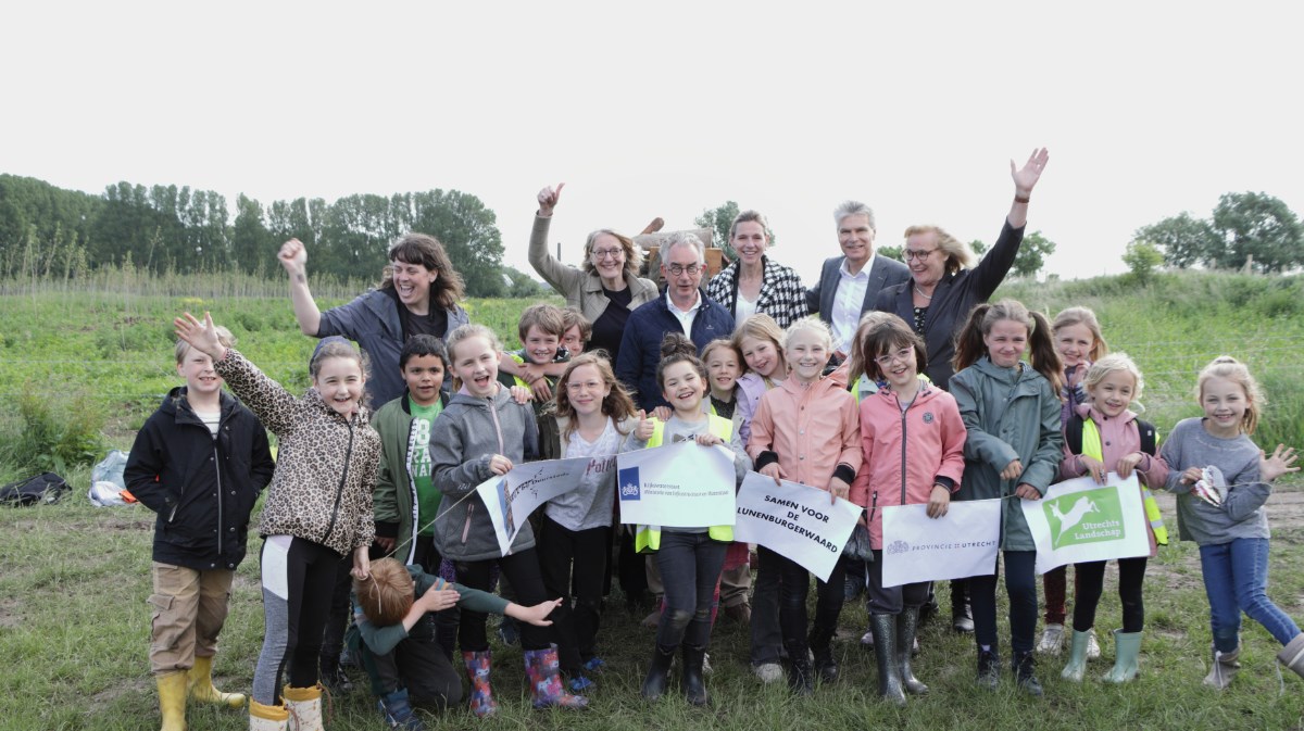 Je ziet een groep kinderen en volwassenen die enthousiast poseren voor een groepsfoto. Ze houden borden vast met teksten die hun steun voor het Lunenburgerwaard-project tonen.